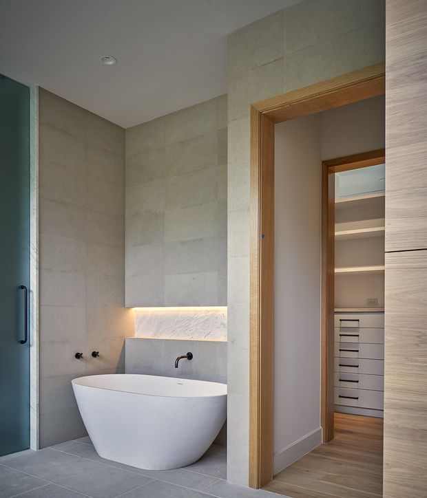 A refined contemporary bathroom featuring a freestanding white bathtub set against large-format pale stone-look wall tiles. A recessed illuminated shelf adds warmth above the tub, with a minimalist dark wall-mounted tap and controls. Timber door frames and a partially visible walk-in wardrobe with built-in drawers and shelving are visible beyond the bathroom.