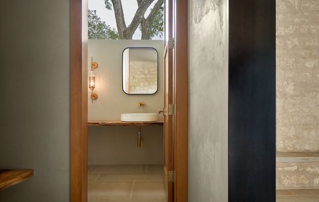 A compact contemporary bathroom viewed through an open timber-framed doorway. The room features a wall-mounted white basin on a thick timber vanity shelf, a brass wall-mounted tap and exposed plumbing, a rounded rectangular mirror, a decorative wall sconce, and large neutral beige floor and wall finishes. Textured plaster walls and dark adjacent wall surfaces add contrast, while a tree is visible through the upper opening.