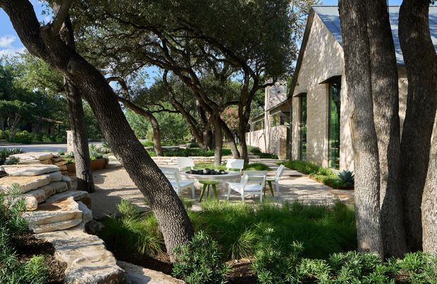 A landscaped outdoor courtyard or terrace beside a stone-clad modern building, shaded by mature spreading trees. The space includes a pale stone-paved seating area with a white outdoor dining set, layered natural-stone retaining steps, low ornamental grasses and shrubs, and tall narrow glazing in the building facade. The setting feels calm, natural, and integrated with the surrounding landscape.