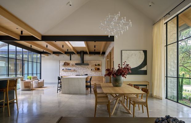 Open-plan contemporary kitchen, dining, and living space with a vaulted ceiling, exposed timber beams, expansive black-framed glazing, polished concrete flooring, and a large kitchen island. The kitchen features light stone surfaces and open shelving, while a sculptural chandelier hangs above the timber dining table.