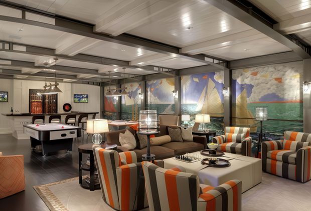 A spacious, richly decorated lounge and entertainment area with exposed dark structural beams, a paneled white ceiling, large nautical mural panels, striped upholstered armchairs and sofas, side tables with lamps, a central coffee table, and a pool table at the rear. The room has a warm, maritime-inspired hospitality or club atmosphere.