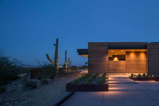 Twilight exterior view of a low, modern flat-roofed residence with dark horizontal cladding, warm recessed lighting, and a concrete entry path. The landscaped frontage includes gravel, raised dark planter beds, drought-tolerant planting, and prominent desert cacti, with distant urban lights and mountains in the background.