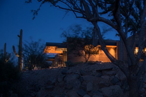 A low, modern home illuminated warmly at dusk, set into a rocky desert landscape with prominent columnar cacti and mature trees. The building has broad horizontal rooflines, extensive glazing, warm wood-toned exterior surfaces, and strong indoor-outdoor visual connections. The dark blue evening sky contrasts with the amber interior and exterior lighting.