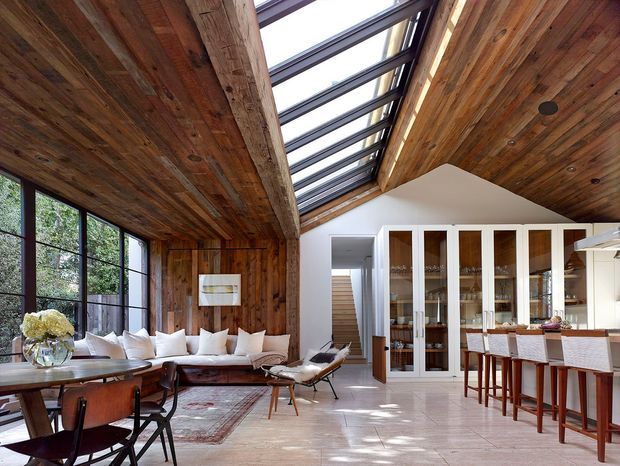 Open-plan interior combining a dining area, living area, and kitchen beneath a dramatic pitched timber ceiling. Warm reclaimed-looking timber boards line the ceiling and walls, with large black-framed glazing and a long central skylight bringing in abundant natural light. A white built-in glass-fronted display cabinet separates the living zone from the kitchen, while a timber dining table, upholstered seating, and a kitchen island with bar stools create a refined contemporary-rustic atmosphere.