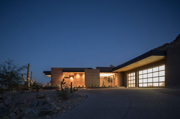 Twilight exterior view of a low-slung contemporary residence with strong horizontal lines, flat roof planes, warm timber-clad walls, and expansive illuminated garage glazing. The home is set within a xeriscaped, rocky desert-style landscape with cacti and a broad paved driveway.