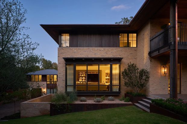 Exterior view of a contemporary two-storey brick house at dusk, set within landscaped gardens. The facade combines warm light brickwork with dark timber upper cladding, broad projecting roof eaves, large illuminated windows, and a covered balcony supported by substantial timber posts. Stepped paths, raised garden beds, low planting, and a small detached illuminated structure are visible around the home.