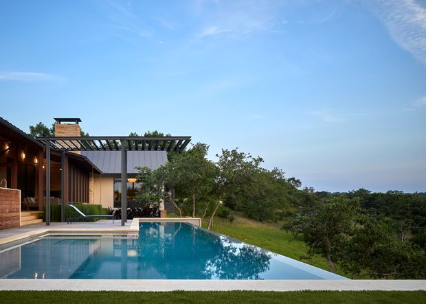 Exterior view of a contemporary residence with a covered outdoor entertaining structure beside an infinity-edge swimming pool. The home uses dark timber cladding and framing with broad glazing, while landscaped grounds extend toward a wooded, elevated outlook under a clear blue sky.