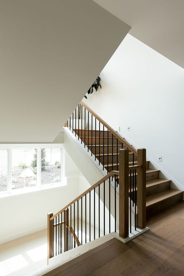 Bright interior view of a multi-level stairway and landing. The staircase has warm timber treads and handrails, dark slender metal balusters, white walls, and large windows that bring in abundant natural light. The open stair void and overlapping angular ceiling planes create a clean, contemporary architectural composition.