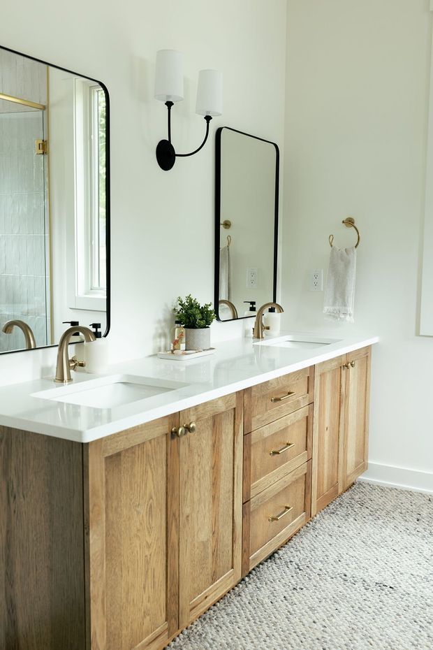 Bright, refined bathroom vanity with a long white double-sink benchtop, warm natural timber cabinetry, brushed brass or gold-toned tapware and hardware, and two black-framed mirrors. White walls, black-and-brass wall sconces, brass towel rings, and a textured pale rug complete the clean, contemporary-traditional look.