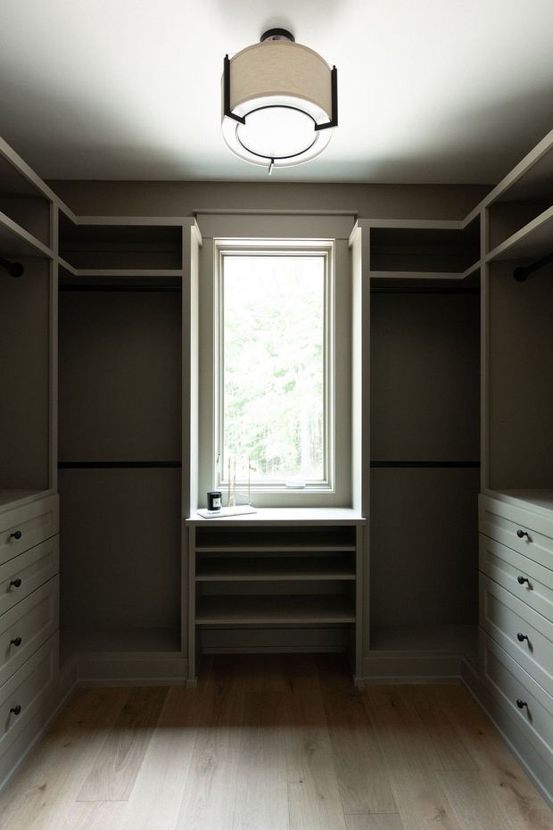 A compact, symmetrical walk-in wardrobe with dark taupe built-in shelving, hanging compartments and white drawer cabinetry on both sides. A central window with a deep sill illuminates the room, while a contemporary circular ceiling light provides additional lighting. The floor is finished in pale natural timber, with simple white trim and a calm, refined palette.