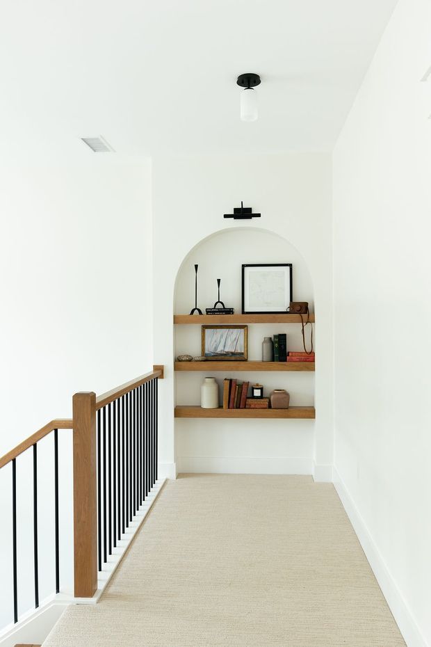 A bright upstairs landing or hallway with white walls and ceiling, a pale carpeted floor, and a recessed arched display niche. Three floating timber shelves hold books, framed artwork, ceramics, and small decorative objects. The landing is bordered by a timber handrail with slim black metal balusters, and minimal black ceiling and wall lighting adds contrast.
