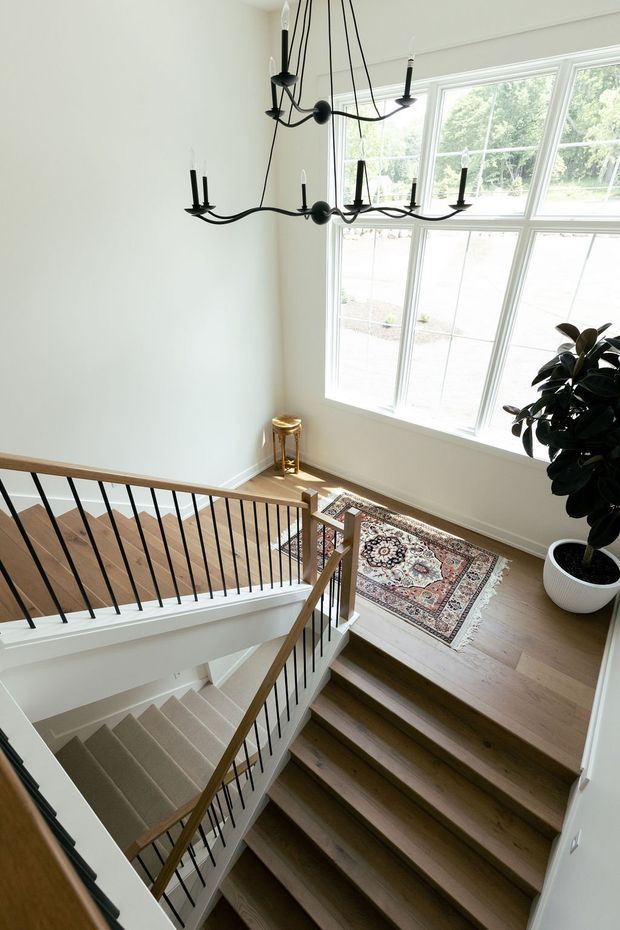 Bright residential stairway and landing viewed from above, featuring two flights of stairs with pale carpeted treads, warm timber flooring and handrails, slim black metal balusters, white walls, a large multi-pane window, a patterned runner rug, a potted rubber plant, and a sculptural black multi-arm chandelier.