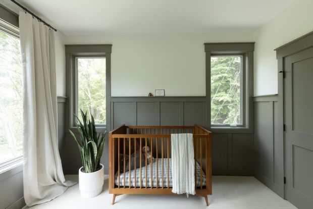 A calm, traditionally detailed nursery with a warm wooden cot centered against dark grey-green painted wall panelling. Tall windows with matching substantial timber trim bring in abundant natural light, while full-length off-white curtains and a potted snake plant soften the room. A small framed print sits above the dado-height panelling, and a paneled door is visible at right.