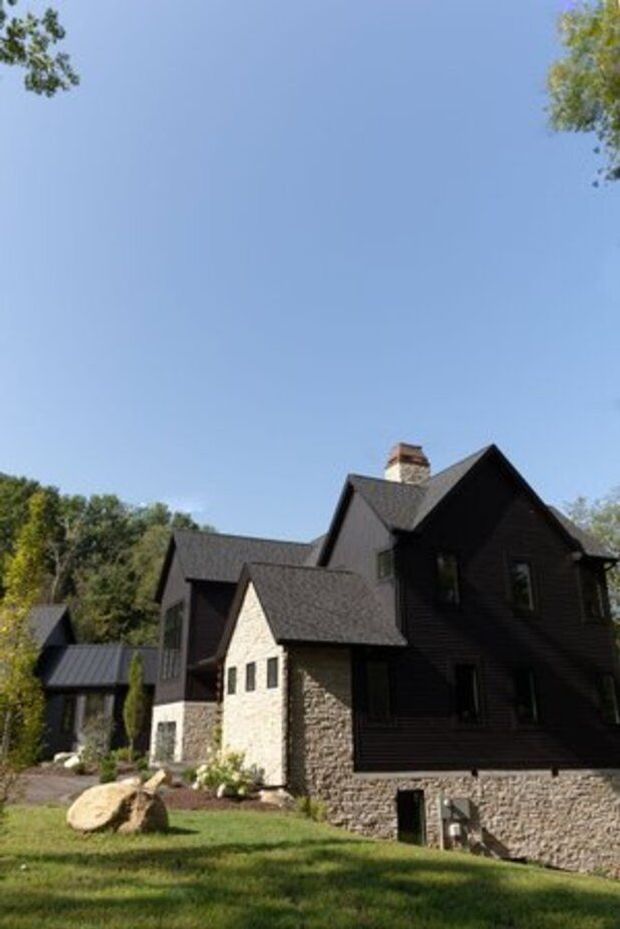Exterior photograph of a large multi-gabled residential house set in a leafy landscaped site. The building combines dark horizontal wood siding, light stone masonry walls and a steep shingled roof with a prominent masonry chimney. A smaller connected wing and covered structure are visible to the left, with lawn, rocks and mature trees surrounding the home.