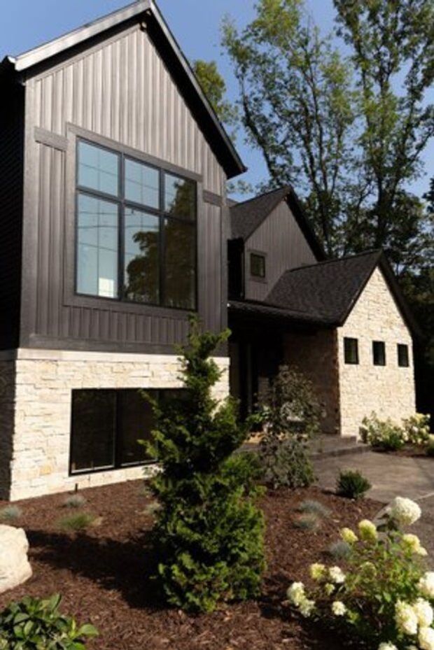Exterior view of a contemporary multi-volume house with steep gabled roofs. The facade combines vertically ribbed grey cladding, pale irregular stone masonry, dark-framed windows, and a landscaped front garden with shrubs and hydrangeas.