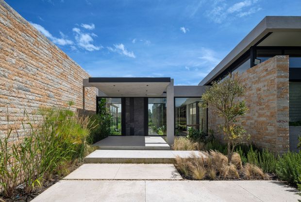 Contemporary single-storey residence viewed from the front approach, with a strongly horizontal composition, flat rooflines, extensive dark-framed glazing, and pale stacked-stone masonry walls. A concrete path and stepped entry platform lead through drought-tolerant ornamental grasses and low planting toward a recessed glass entrance. The image is an exterior architectural photograph under a bright blue sky.