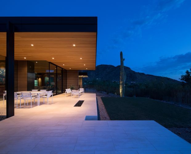 Dusk exterior view of a contemporary flat-roofed house with extensive floor-to-ceiling glazing, a deep timber-lined soffit, dark structural framing, and a broad pale paved terrace furnished for outdoor dining and lounging. The property opens toward a landscaped yard and rugged mountain backdrop, with integrated recessed lighting creating a warm glow.