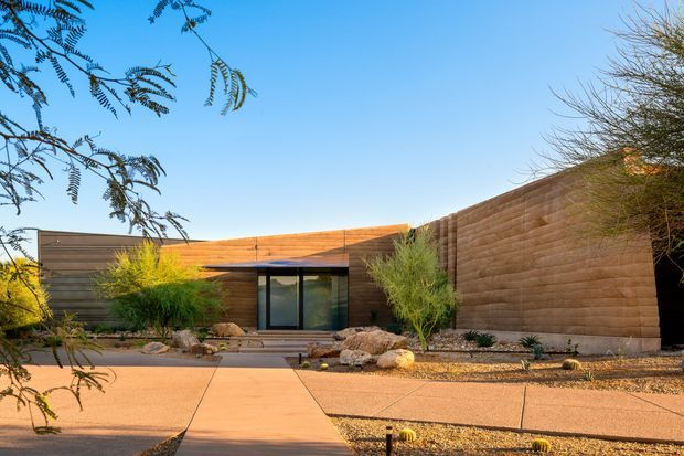 Single-storey contemporary residence with a low, horizontally composed form and warm rust-brown rendered walls. A dark-framed glazed entry sits beneath a projecting roof, opening onto a broad concrete path. The landscaped front garden uses gravel, desert planting, sculptural rocks, and small trees, creating a restrained Southwestern-style courtyard approach under a clear blue sky.