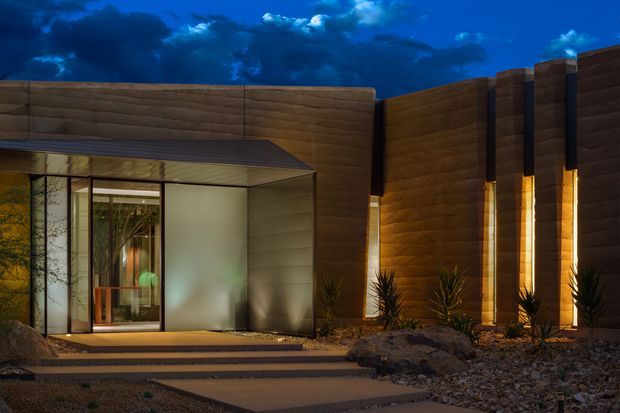Dusk exterior view of a contemporary single-storey building with dark horizontal cladding, a projecting flat-roofed glazed entry pavilion, tall illuminated wall sections, and a minimalist gravel landscape with architectural planting and stepping slabs.