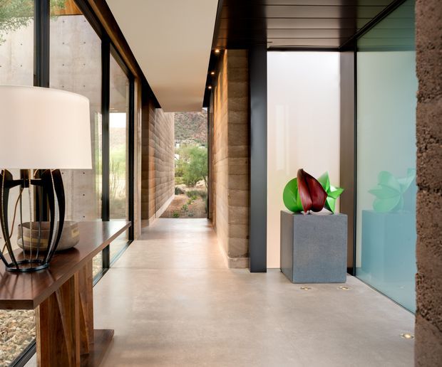 A refined modern interior corridor or entry passage with a polished concrete floor, exposed textured masonry walls, extensive black-framed glazing, and a dark timber-lined ceiling. A timber console and sculptural lamp sit beside the glazing, while a minimalist pedestal and abstract plant-like sculpture form a focal point near frosted glass doors. The passage opens toward a landscaped exterior, creating a strong indoor-outdoor connection.