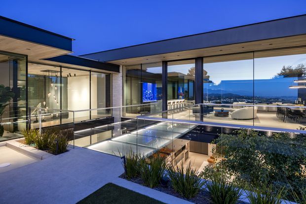 A luxurious contemporary multi-level residence at dusk, arranged around a sunken central courtyard or pool-like glazed void. Extensive floor-to-ceiling glazing, flat roof planes, dark metal framing, pale stone or concrete terraces, and warm integrated lighting create a seamless indoor-outdoor composition. The living area is visible through the glazing, with a distant mountainous waterfront landscape beyond.