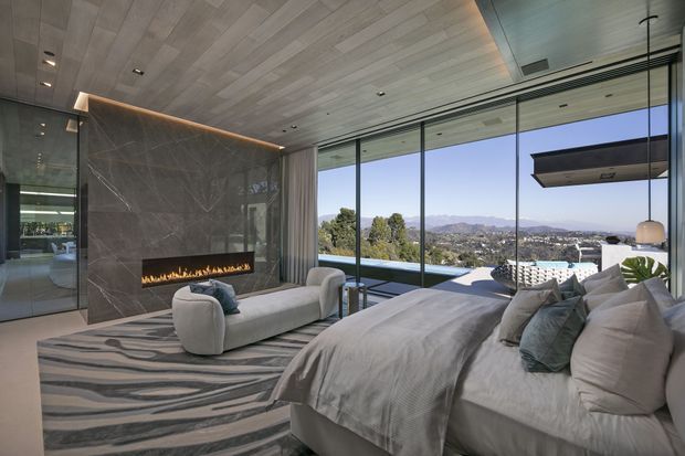 Luxurious contemporary bedroom with a partially visible living area, featuring a large upholstered bed, sculptural lounge seating, a linear built-in fireplace set into a dark stone feature wall, full-height glazing, and a panoramic urban hillside view. The ceiling and feature wall use refined, large-format finishes, while integrated lighting and a suspended fireplace hood create a polished high-end atmosphere.