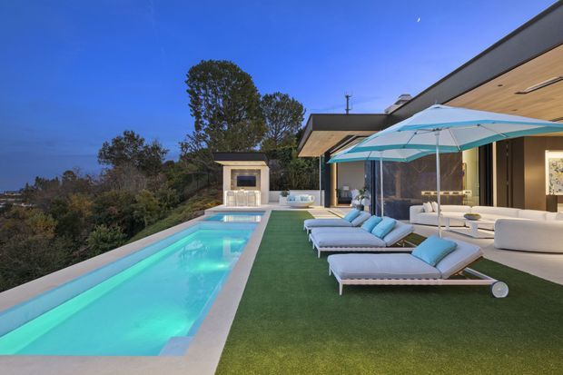 Twilight view of a modern residential pool terrace on a landscaped, elevated site. A long turquoise swimming pool runs alongside a broad artificial-grass lounging area furnished with pale upholstered sun loungers and a large white-and-turquoise umbrella. The adjoining contemporary house has dark-framed glazing, a deep roof overhang with timber-lined soffit, and an outdoor fireplace pavilion at the far end. The setting feels refined, relaxed, and resort-like.
