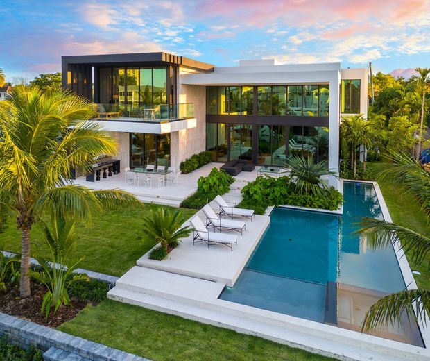 Aerial exterior view of a large contemporary two-storey residence with crisp white and dark charcoal rectilinear volumes, extensive floor-to-ceiling glazing, balconies, and landscaped tropical gardens. A long geometric swimming pool with adjoining pale paved terraces dominates the foreground, while palm trees surround the property.