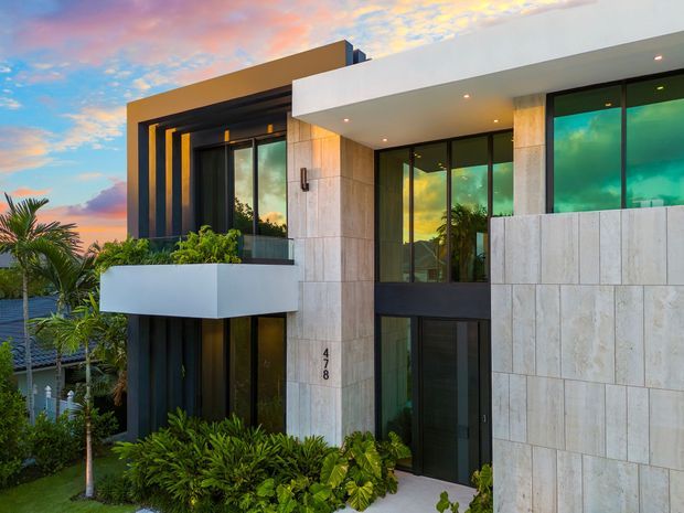 Exterior view of a contemporary multi-storey residence at sunset, featuring a flat roof, large dark-framed floor-to-ceiling glazing, projecting white balcony, dark vertical facade elements, and pale stone wall cladding. Lush tropical landscaping surrounds the entrance and balcony.