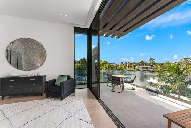 A bright contemporary interior opening onto a waterfront terrace. The room features white walls, a dark low dresser, black armchair, pale timber flooring, a large light rug, and a round textured wall mirror. Full-height black-framed glazing and a folding glass door lead to a covered terrace with outdoor seating, overlooking a calm waterway and palm-lined surroundings. Exposed timber pergola beams provide shade above the terrace.
