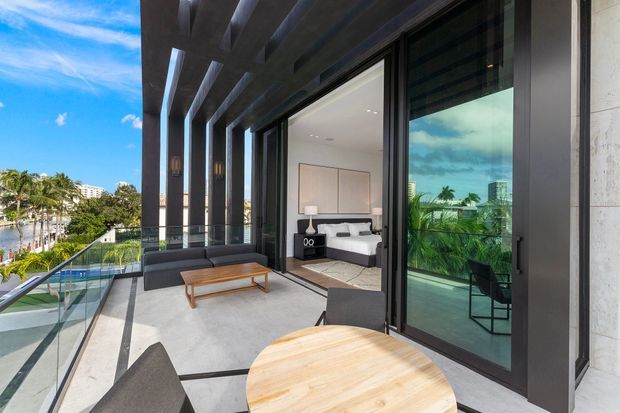 A bright contemporary balcony terrace with dark structural framing, deep overhang beams, glass balustrades and large sliding glass doors. Outdoor seating and timber tables overlook a lush landscaped setting and pool, while the bedroom interior is visible through the open glazing. The space has a refined indoor-outdoor character with strong daylight and tropical greenery beyond.