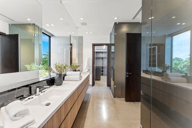 Bright contemporary bathroom with a long double vanity, white integrated basins, warm timber cabinetry, large mirrors, pale stone-look flooring, glass shower enclosure, and dark timber doors. A freestanding bathtub is visible through the glass on the right, with natural greenery and views beyond the windows.