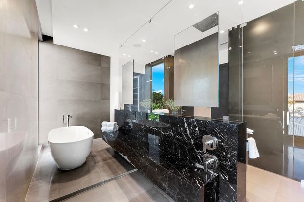Luxurious contemporary bathroom with a freestanding white bathtub, large-format pale stone wall tiles, warm-toned floor tiles, and a dramatic black marble double vanity with integrated basins and chrome fixtures. Full-height mirrors and glazing reflect the bright, open space, while recessed ceiling lighting creates an airy, refined atmosphere.