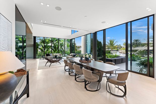A bright, spacious contemporary open-plan interior combining a dining area and living room. A long sculptural dining table with upholstered chairs occupies the foreground, while a low sofa and lounge seating sit beyond it. Full-height black-framed glazing wraps the room, opening views to lush tropical landscaping, palm trees, blue sky, and neighbouring homes. The space has pale timber or stone-look flooring, white walls and ceiling, restrained furnishings, and abundant natural light.