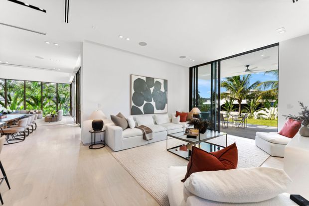 Bright, spacious contemporary living room with white walls, pale timber flooring, floor-to-ceiling black-framed glazing, and an open connection to a lush tropical outdoor area. The room contains a large white sofa, sculptural lounge chairs, a glass coffee table, neutral textured rug, and rust-coloured accent cushions. A dining or secondary sitting area is visible to the left, while the main focus is the refined indoor-outdoor living space.