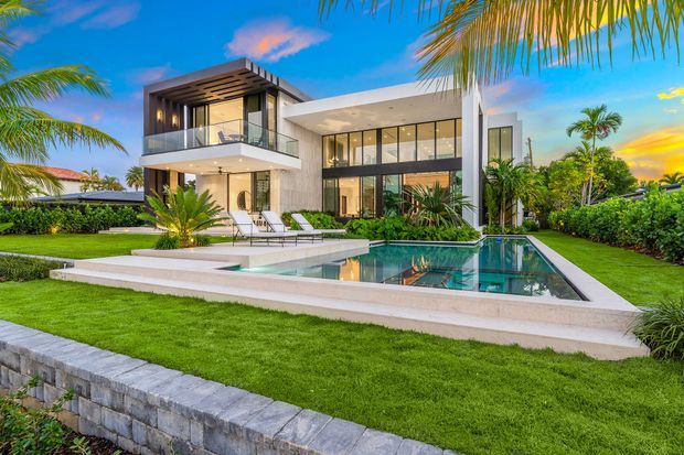 Exterior view of a large contemporary two-storey residence with extensive floor-to-ceiling glazing, white and grey facade surfaces, a projecting upper-level balcony, and a flat roof. A geometric swimming pool with a raised white-edged terrace occupies the foreground, surrounded by manicured lawn, palms, and hedges.
