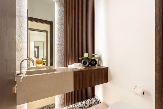 A refined contemporary bathroom featuring a wall-mounted stone vanity with integrated rectangular basin and brushed metal tapware. Dark timber cabinetry and vertical timber battens create a warm feature wall, complemented by a large framed mirror, pebble-filled floor detail, and minimal white walls. A toilet is partially visible at right.