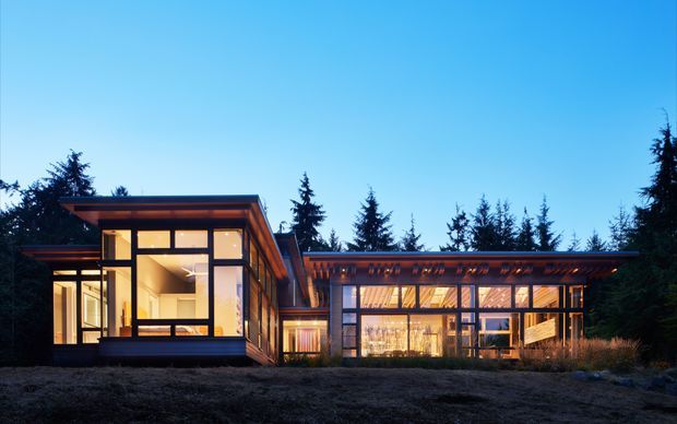 A modern single-storey residence photographed at dusk, set among dense evergreen trees. The building has a low, horizontally composed roofline, extensive floor-to-ceiling glazing, dark structural framing, warm timber-toned cladding, and a bright glazed living area. A projecting glazed wing and sheltered central section create a linear pavilion-like form.