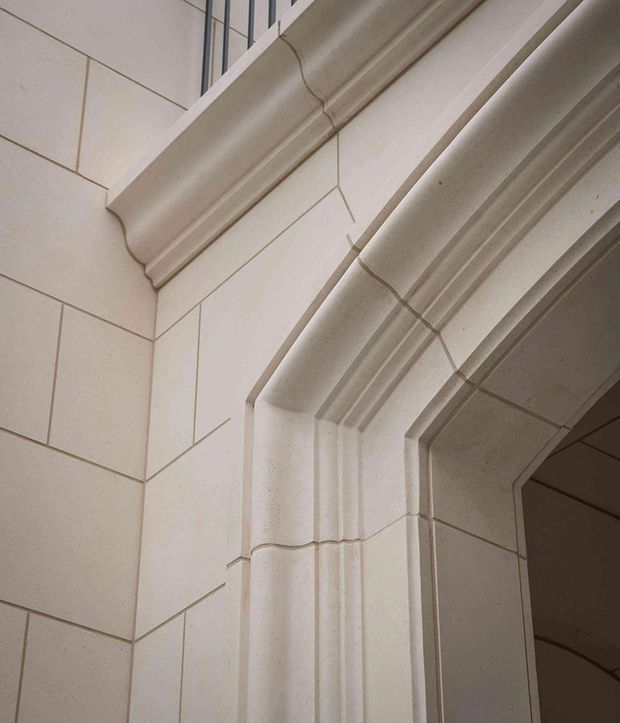 Close-up architectural detail of a pale stone-clad wall and deeply moulded, stepped archway or doorway surround. Large rectangular stone panels are arranged with fine recessed joints, while layered curved and vertical trim creates a formal, highly articulated opening. A darker interior space is partially visible beyond the opening.