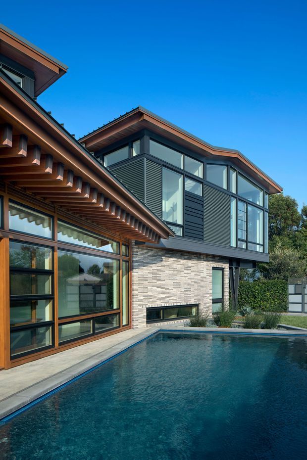 Exterior view of a contemporary multi-level residence beside a swimming pool. The building combines extensive dark-framed glazing, horizontal dark cladding, warm timber eaves and soffits, and a light textured brick wall. The roof forms are low-pitched with broad projecting eaves and exposed timber detailing. Landscaped hedging and the pool surround the house, under a clear blue sky.