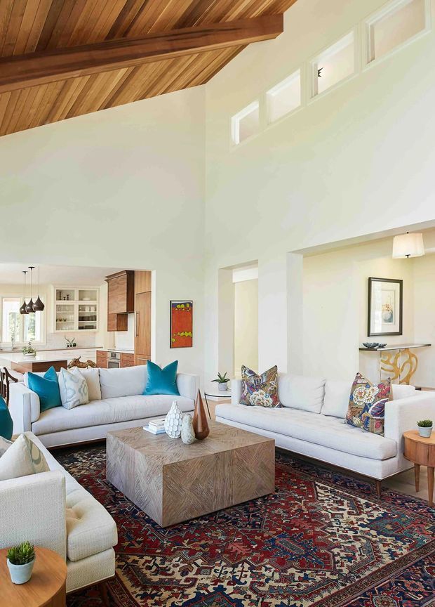 Bright, spacious living room with white walls and high ceilings, furnished with several light upholstered sofas, a large patterned Persian-style rug, and a substantial square timber coffee table. A richly stained timber vaulted ceiling with exposed beams dominates the upper frame. The room opens toward a white and timber kitchen at the rear, with high clerestory windows, colourful patterned cushions, a decorative fireplace mantel, and abundant natural light.