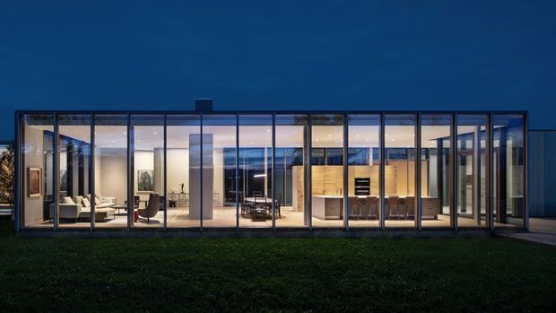 A low, single-storey modern residential building photographed at dusk, with an extensive floor-to-ceiling glazed facade framed in dark metal. Warm interior lighting reveals an open-plan living area, dining space, and kitchen with timber-toned cabinetry, while a dark flat roof and broad lawn reinforce the minimalist horizontal composition.