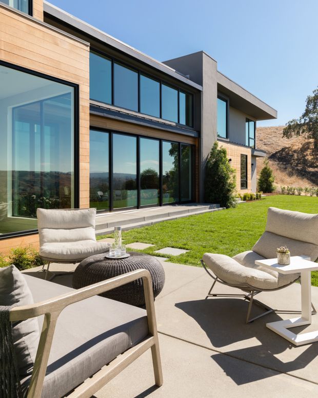 Bright exterior view of a contemporary multi-level residence with expansive floor-to-ceiling glazing, warm horizontal timber cladding, and smooth concrete or rendered walls. A paved outdoor sitting area with neutral lounge chairs and a textured round coffee table overlooks a manicured lawn and dry, gently sloping landscape. The scene has strong sunlight, clean lines, and a seamless indoor-outdoor connection.