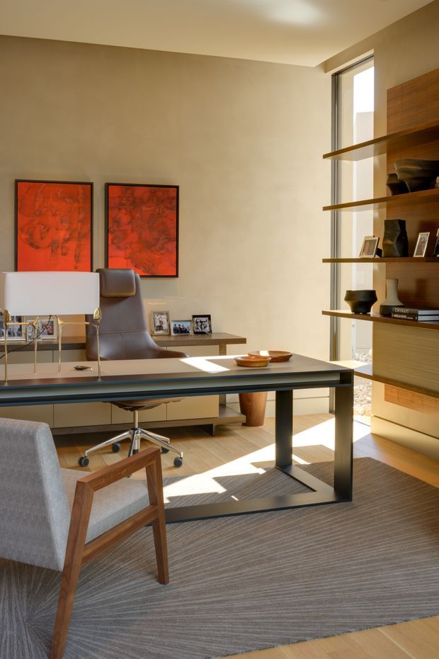 A refined modern home office with a large dark-framed desk, contemporary task chair, upholstered guest chair, and table lamp. The room features warm beige plaster walls, light timber flooring, a grey textured area rug, expansive vertical glazing, and a built-in timber shelving unit displaying books and decorative objects. Two vivid red abstract artworks provide a strong accent against the neutral interior.