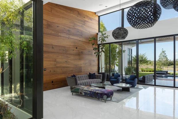 A bright, high-volume contemporary living room with floor-to-ceiling black-framed glazing, a dramatic full-height timber feature wall, polished pale flooring, and views to a landscaped outdoor area and pool. The room is furnished with a grey sofa, sculptural blue lounge chairs, a patterned low ottoman, and large perforated spherical pendant lights.