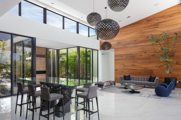A bright, contemporary open-plan living and dining space with a double-height ceiling and extensive black-framed glazing. A long marble or pale stone island with four upholstered bar stools separates the kitchen area from the lounge. The living zone includes a grey sofa, blue lounge chairs, a low coffee table and a large pale rug. A dramatic full-height timber feature wall and clustered perforated spherical pendant lights add warmth and visual interest, while lush greenery is visible through the glazing.
