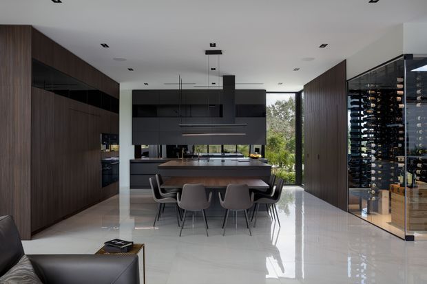 A spacious contemporary open-plan kitchen and dining area with dark timber cabinetry, matte black overhead joinery, a central island with integrated dining table, and six modern dining chairs. A full-height glass-enclosed wine display is visible on the right, while large glazing at the rear opens views to greenery. Polished light flooring, recessed lighting, and clean linear forms create a refined, high-contrast interior.