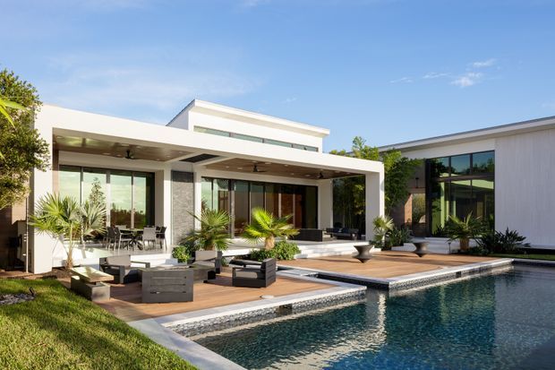 Contemporary single-storey residence with crisp white rendered volumes, extensive floor-to-ceiling glazing, and a covered outdoor entertaining area opening onto a timber-look deck and rectangular swimming pool. The landscaped courtyard includes palms, built-in seating, and outdoor lounge furniture, creating a bright, resort-like setting.