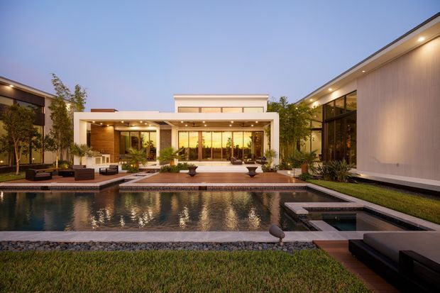 A contemporary single-storey residence arranged around a landscaped courtyard and rectangular swimming pool. The dusk view shows flat rooflines, extensive floor-to-ceiling glazing, pale rendered walls, warm timber-toned vertical cladding, deep covered outdoor living areas, and integrated architectural lighting. Poolside paving, planting, dark stone edging, and outdoor lounge furniture frame the reflective water feature.