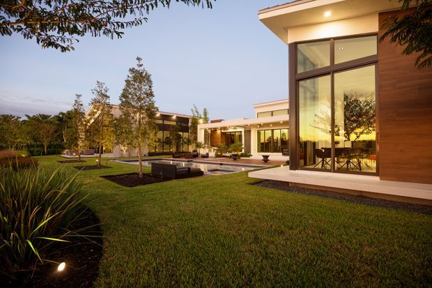 A modern low-profile residence viewed from a landscaped lawn at dusk. The home features expansive floor-to-ceiling glazing, warm timber wall cladding, flat rooflines, broad eaves, and a connected courtyard with paving, planting, seating, and a narrow reflecting pool or water feature. Interior dining and living areas are visible through the glass. The scene has carefully integrated architectural lighting and a refined contemporary outdoor setting.